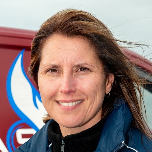 woman with brown hair wearing a blue jacket, standing in front of a red sign with a blue flame symbol